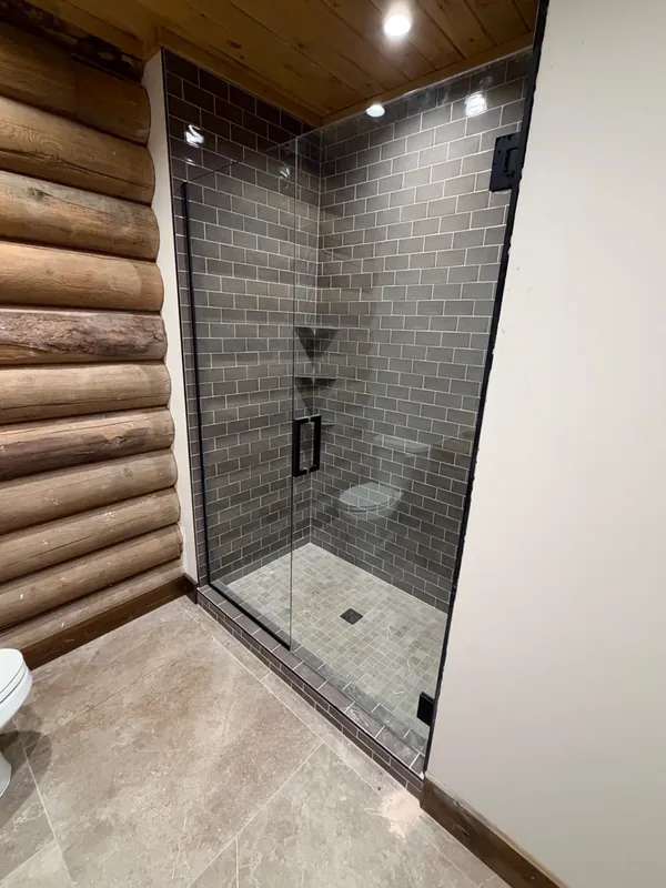 Island Park cabin: Frameless glass shower with matte black hinges and pull handle with gray subway tile and log walls