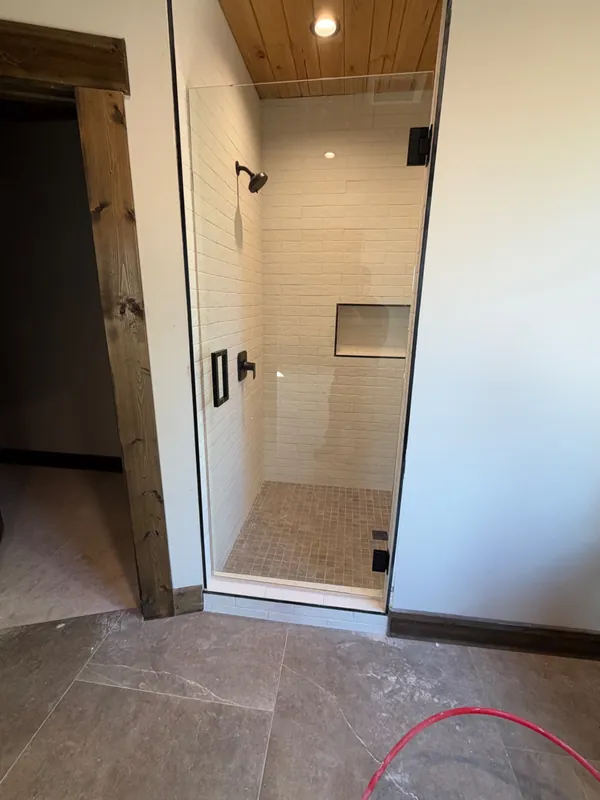 Island Park cabin: Single frameless glass shower door with matte black hinges and handle under a wood plank ceiling
