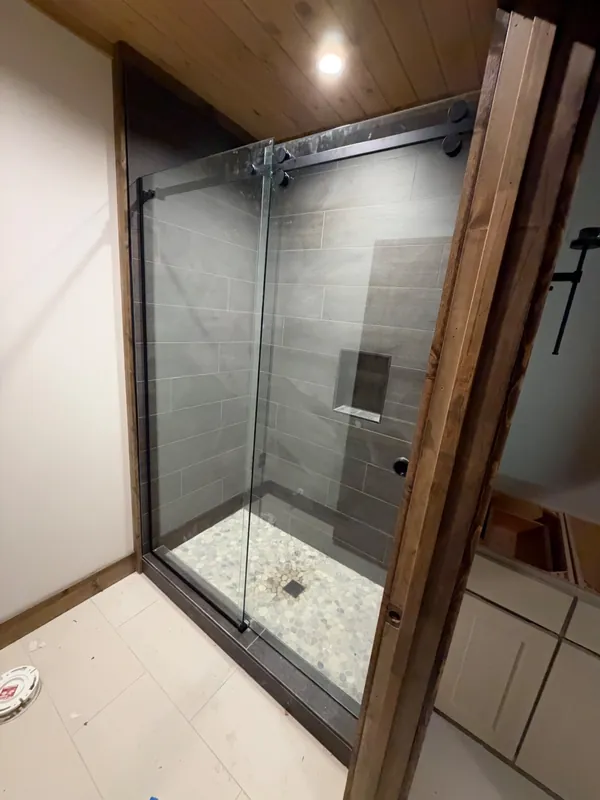 Island Park cabin: Frameless sliding glass shower door on a matte black roller track with dark plank tile and pebble floor