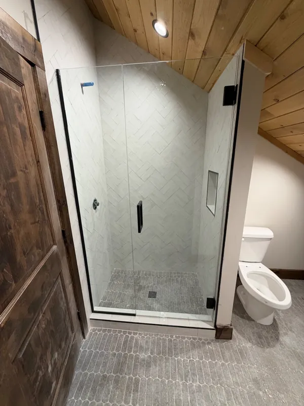 Island Park cabin: Frameless shower door and panel under a sloped wood ceiling with white herringbone tile and matte black hardware