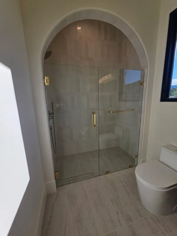 Teton Valley home: Frameless glass shower door and panel in a tiled arched alcove with brushed gold hinges, clamps, and pull handle